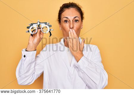 Middle age senior oculist woman holding optometrical glasses for vision correction cover mouth with hand shocked with shame for mistake, expression of fear, scared in silence, secret concept
