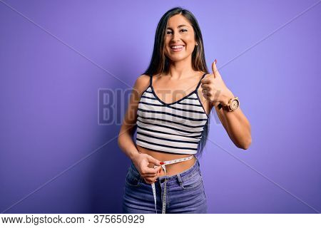 Young beautiful woman using measure tape on waist looking for healthy slim body happy with big smile doing ok sign, thumb up with fingers, excellent sign