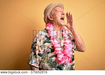 Grey haired senior man wearing summer hat and hawaiian lei over yellow background shouting and screaming loud to side with hand on mouth. Communication concept.