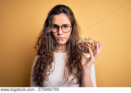 Beautiful woman with curly hair holding bowl with healthy peanuts over yellow background with a confident expression on smart face thinking serious