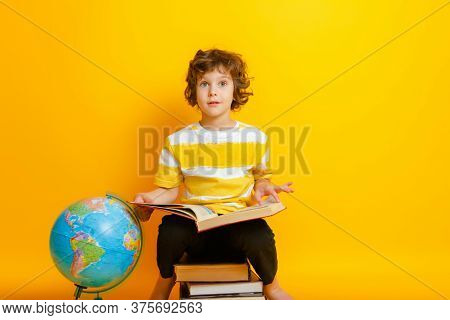 Curly Boy In Glasses And Stripes Tshirt And Sits On A Stack Of Books, Near To The Globus On Yellow B