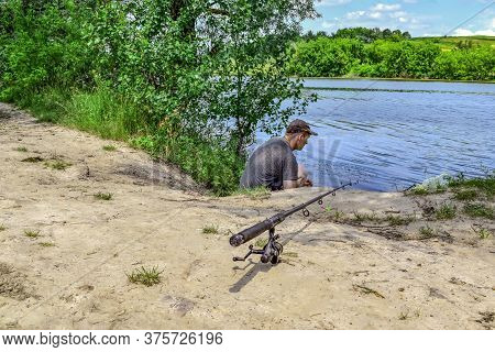 Fishing Rod Stands On The Ground Close-up Against Of A Young Adult Man Near The Pond On A Sunny Summ