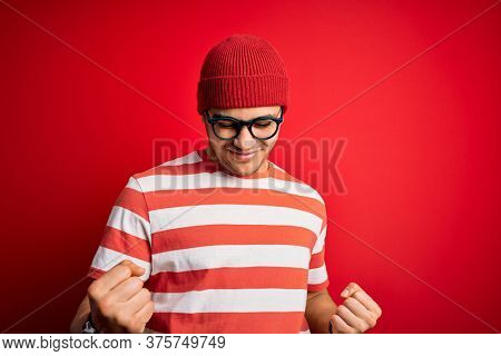 Young brazilian man wearing casual striped t-shirt and glasses over isolated red background very happy and excited doing winner gesture with arms raised, smiling and screaming for success. Celebratio