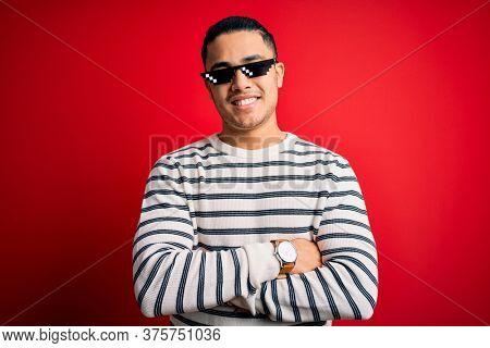 Young brazilian man wearing funny thug life sunglasses over isolated red background happy face smiling with crossed arms looking at the camera. Positive person.