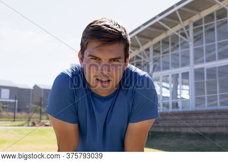 Portrait close up of a Caucasian male runner training at a sports field, wearing a blue t shirt, looking to camera and leaning forward recovering after a run
