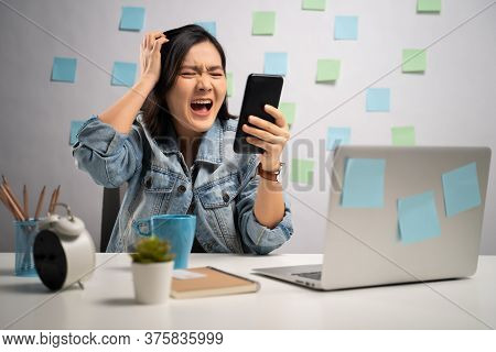 Asian Woman Angry Looking At Smart Phone Reading News And Shouting At Home Office. Wfh. Work From Ho