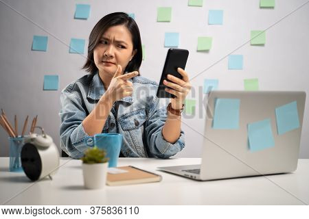 Asian Woman Angry Looking At Smart Phone Reading News At Home Office. Wfh. Work From Home. Preventio