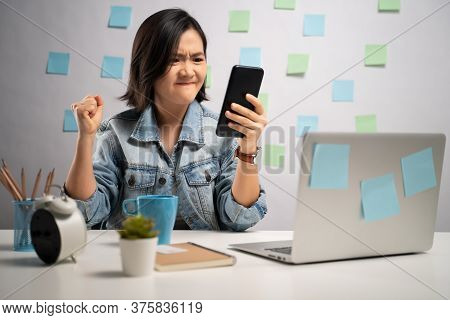 Asian Woman Angry Looking At Smart Phone Reading News At Home Office. Wfh. Work From Home. Preventio