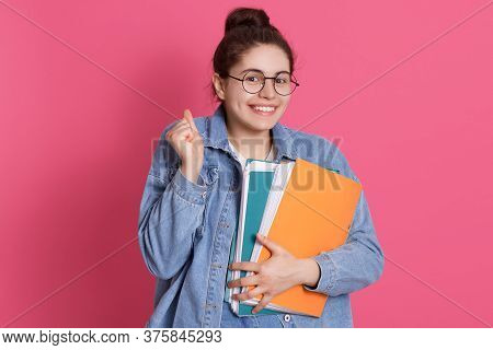 Young Smiling Student In Eyeglasses And Denim Jacket Standing With Paper Folders Isolated Over Rosy 