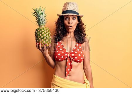 Middle age beautiful woman wearing bikini and summer hat holding pineappel scared and amazed with open mouth for surprise, disbelief face 