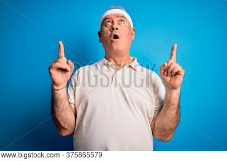 Middle age hoary sportsman doing sport wearing sportswear over isolated blue background amazed and surprised looking up and pointing with fingers and raised arms.
