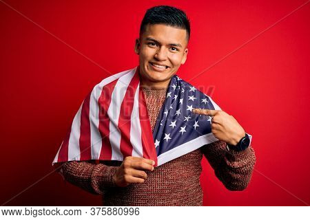 Young handsome latin patriotic man wearing united states of america falg very happy pointing with hand and finger