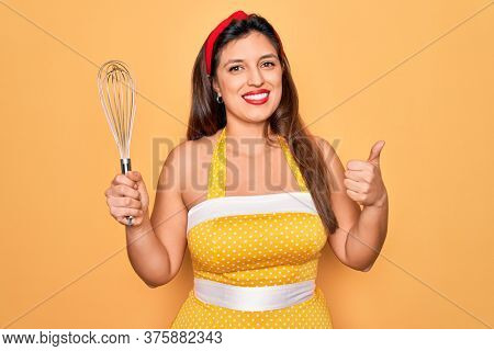 Young hispanic pin up woman wearing fashion sexy 50s style holding cooking whisk blender happy with big smile doing ok sign, thumb up with fingers, excellent sign