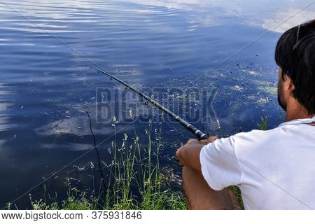 Fisherman With Fishing Rod At The Pond. A Man In A White T-shirt Is Fishing Against A Background Of 