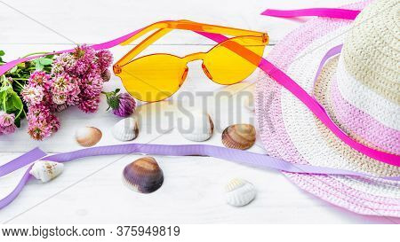 Yellow Rimless Glasses Between A Straw Hat And Pink Flowers On A White Background Among Seashells. T