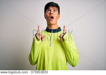 Young handsome sportsman doing sport wearing sportswear over isolated white background amazed and surprised looking up and pointing with fingers and raised arms.