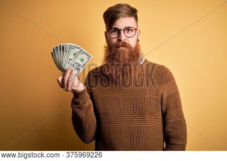Redhead Irish man with beard holding a bunch of 1 dolla banknotes over yellow background with a confident expression on smart face thinking serious