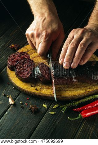 Chef's Hands With A Knife Cut Blood Sausage On A Kitchen Cutting Board. Cooking A National Dish With