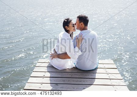 Young Couple In Love In Light Casual Clothes Is Sitting On Wooden Bridge. Happy Woman And Man Hug An