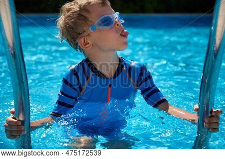 Funny Boy In A Blue Bathing Suit Stands On A Step In The Pool, Holding On To A Metal Railing, Shows 
