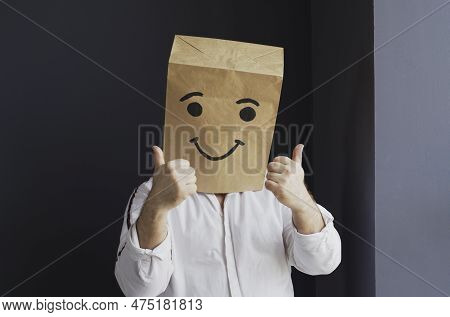 A Man In A White Shirt With A Paper Bag On His Head, With A Smiley Face Drawn, Stands Against The Wa