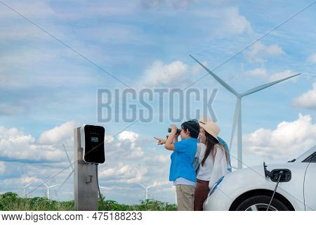 Concept Of Progressive Happy Family Enjoying Their Time At Wind Farm With Electric Vehicle. Electric