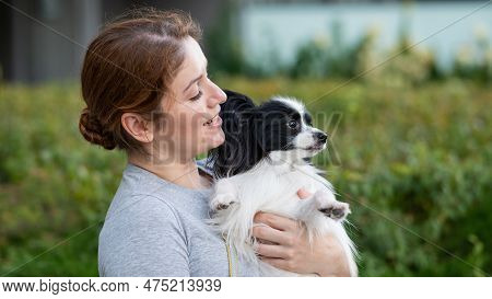 Caucasian Red-haired Woman Holding Pappilion Dog In Her Arms Outdoors. Black And White Continental S