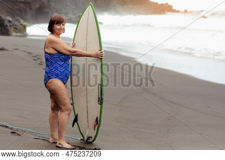 Joyful Adult Middle Aged Woman In Swimsuit Holds Surfboard To Go Surfing, Enjoys Summer Vacation At 