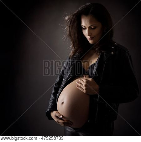 Stylish Pregnant Woman In Biker Leather Jacket Hugging Abdomen. Pregnancy Belly Side View On Dark Ba