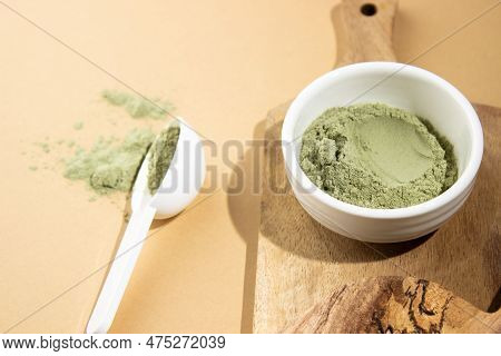 Green Powder In A Small White Ceramic Bowl, With A White Spoon On A Wooden Board, On A Beige Backgro