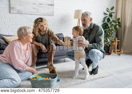 Cheerful Bearded Man Supporting Baby Girl Doing Her First Steps Near Family In Living Room.
