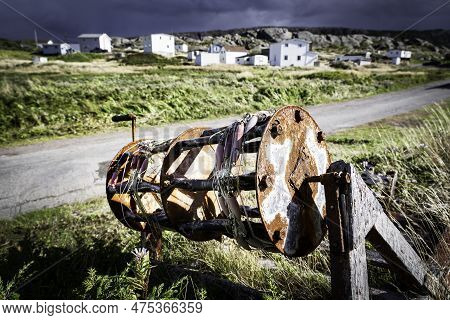 Rustic Hand Reel From A Commercial Fishing Boat Lying In Tall Grass Overlooking Small Wooden Homes A