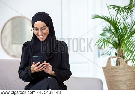 Muslim Young Woman In Black Hijab And Burqa Sitting On Sofa At Home And Using Mobile Phone Smiling.