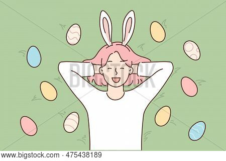Funny Girl With Rabbit Ears Lies On Grass Among Colorful Easter Eggs And Smiles Broadly. Carefree Fe