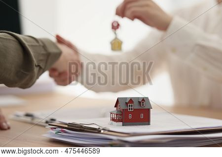 Female Real Estate Agent Gives Key To House To Client Shaking Hand. Small Model Of Townhouse Stands 