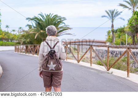 Rear View Of Senior Gray Haired Woman With Backpack Walking In Public Park At Sea Enjoying Retiremen