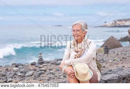Portrait Of Pensive Caucasian Woman Traveler Sitting At The Beach Holding Backpack. Smiling Elderly 