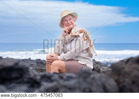 Portrait Of Smiling Senior Woman Wearing Eyeglasses And Summer Hat Sitting Outdoors At Sea Enjoying 