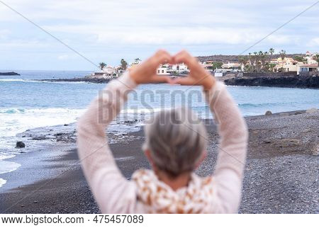 Rear View Of Defocused Senior Woman Standing Outdoors At Sea Looking At Horizon Gesturing Heart Shap