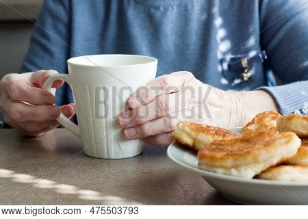 Grandmother Eating Pancakes And Drinking Tea Sitting At Kitchen Table In House. Closeup Of Wrinkled 