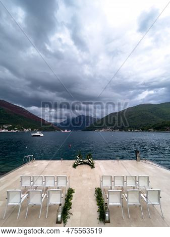 Rows Of White Chairs Stand In Front Of A Wedding Semi-arch On A Pier In The Sea Overlooking A Mounta