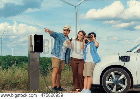 Concept Of Progressive Happy Family Enjoying Their Time At Wind Farm With Electric Vehicle. Electric