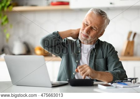 Senior Man Suffering Neckache While Working With Laptop In Kitchen Interior