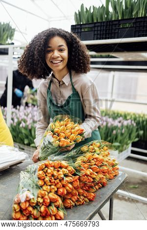 Cute Attractive African Girl In Green Apron Holding Bouquet Of Orange Tulips Indoors In Greenhouse. 
