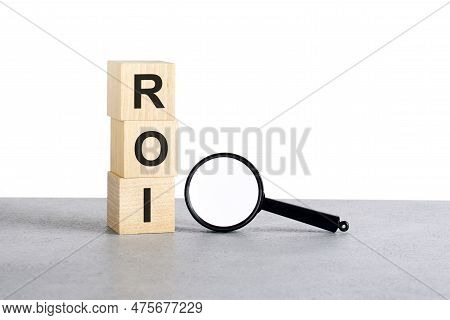 Roi - Return On Investment - Text On Wooden Cube Blocks And Magnifying Glass On Grey Table