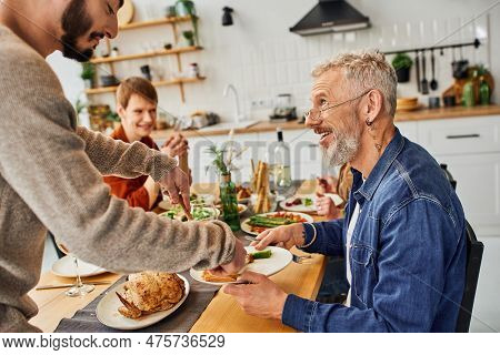 Smiling Bearded Man Looking At Gay Man Serving Chicken During Supper With Family In Kitchen.