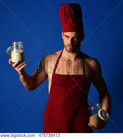 Serious Male Chef In Burgundy Hat And Apron Holds Glass Jars With Groats And Cereals. Professional C