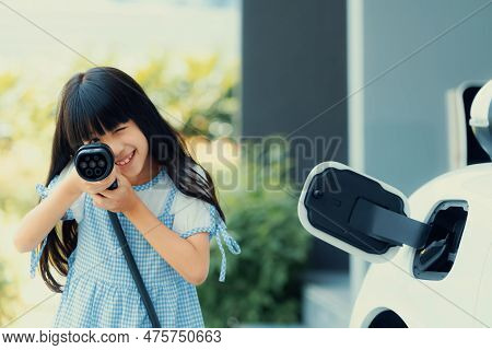 A Playful Girl Holding And Pointing An Ev Plug, A Home Charging Station Providing A Sustainable Powe
