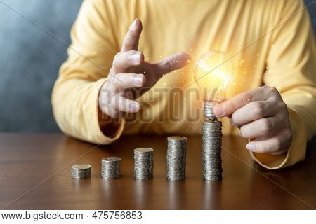 Man Holding A Glowing Abstract Light Bulb With Pile Money Cions On A Wooden Table, Business Problem 