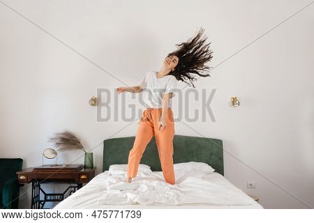 Happy Young Caucasian Woman Dancing On Bed In Bedroom. Dark Hair Fly Away. Freedom And Relax At Home